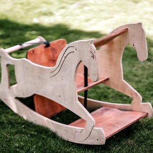 May include: A wooden rocking horse with a light brown finish, set on a green grassy surface. The horse features a detailed head and body design, with a red-brown seat and base. The rocking horse is designed for children.
