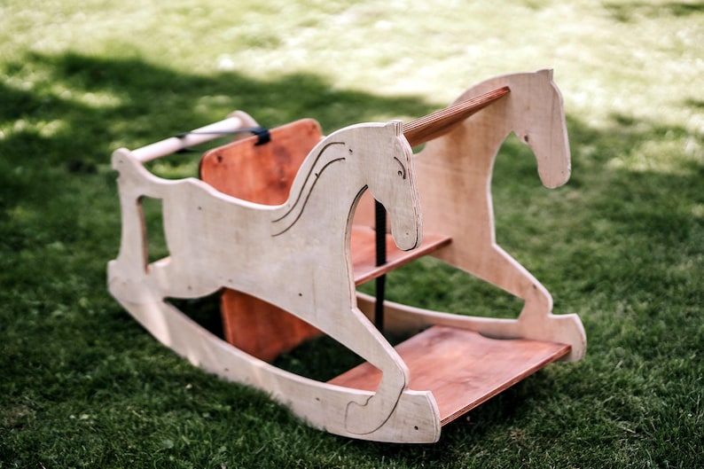 May include: A wooden rocking horse with a light brown finish, set on a green grassy surface. The horse features a detailed head and body design, with a red-brown seat and base. The rocking horse is designed for children.