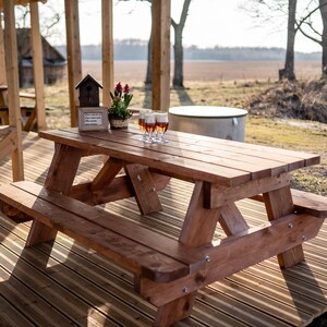 May include: A wooden picnic table with a bench on a wooden deck. There are four glasses of beer on the table, a birdhouse, and a potted plant with red flowers.