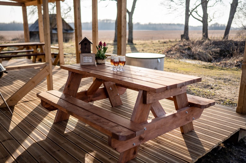 May include: A wooden picnic table with a bench on a wooden deck. There are four glasses of beer on the table, a birdhouse, and a potted plant with red flowers.
