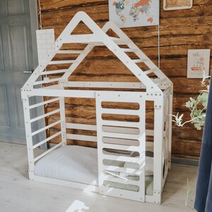 May include: White wooden playhouse with a ladder and a bed inside. The playhouse is designed to look like a house with a roof and windows. The playhouse is in a room with a wooden wall and a door.