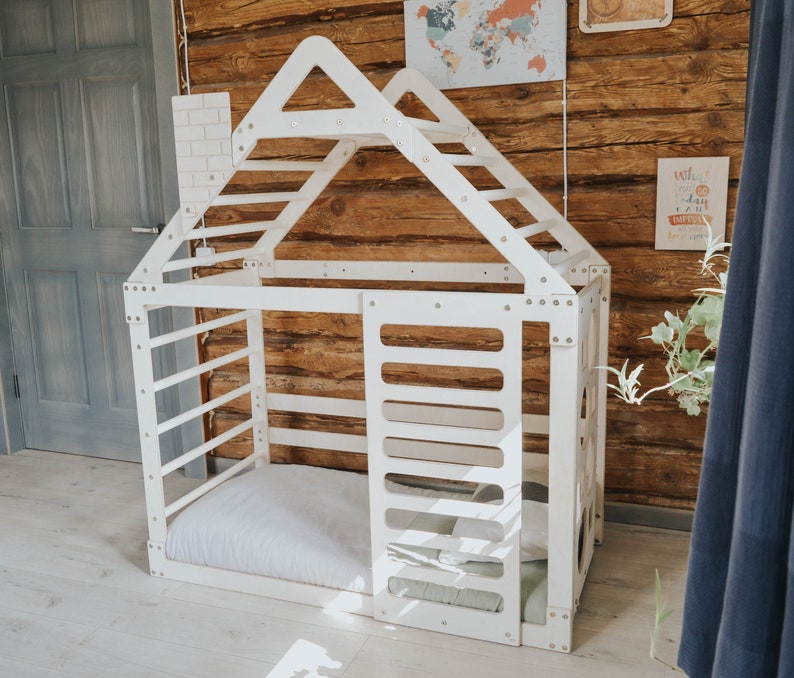 May include: White wooden playhouse with a ladder and a bed inside. The playhouse is designed to look like a house with a roof and windows. The playhouse is in a room with a wooden wall and a door.