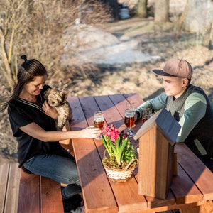 May include: A couple sits at a wooden picnic table outdoors with a small dog. The woman is holding the dog and the man is looking down. There are two glasses of beer on the table and a potted plant with pink flowers.