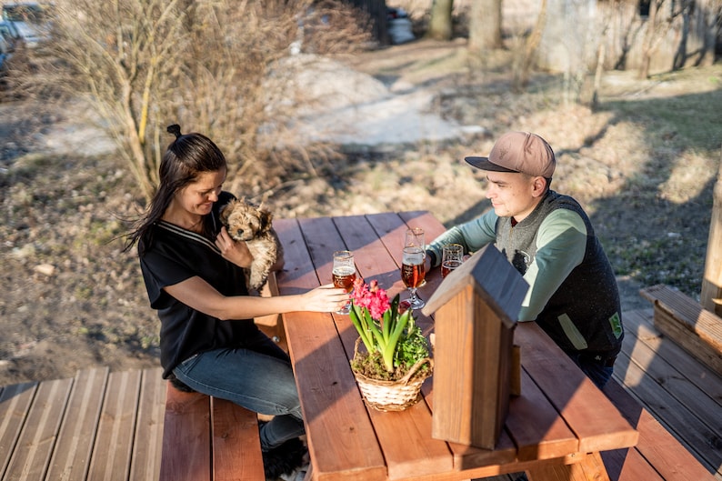 May include: A couple sits at a wooden picnic table outdoors with a small dog. The woman is holding the dog and the man is looking down. There are two glasses of beer on the table and a potted plant with pink flowers.