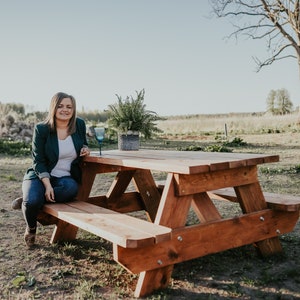 May include: A woman sits on a wooden picnic table with a glass of water. The table is made of natural wood and has a rustic look. The woman is wearing a green blazer and jeans. The table is set in a grassy field with trees in the background.