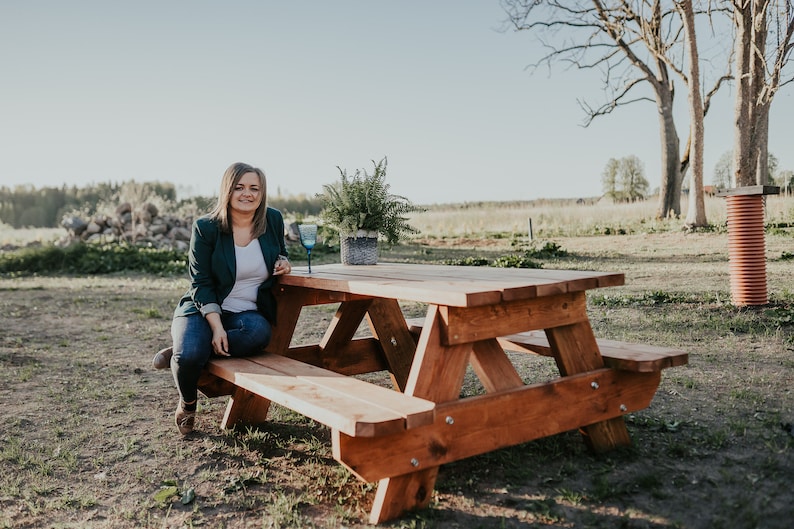 May include: A woman sits on a wooden picnic table with a glass of water. The table is made of natural wood and has a rustic look. The woman is wearing a green blazer and jeans. The table is set in a grassy field with trees in the background.