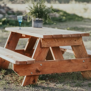 May include: A wooden picnic table with two benches, made of natural wood with a reddish-brown stain. A blue glass sits on the table.
