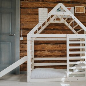 May include: White wooden playhouse with a slide, a climbing wall, and a ladder. The playhouse has a bed inside and is set against a wooden wall.