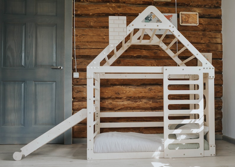 May include: White wooden playhouse with a slide, a climbing wall, and a ladder. The playhouse has a bed inside and is set against a wooden wall.