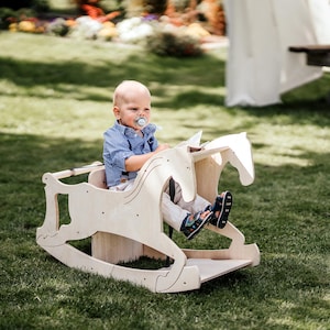 May include: A light-colored wooden rocking horse with a child seated in it, positioned on a grassy lawn. The horse features a cutout design and a seat. The child is wearing a blue shirt and light-colored pants.