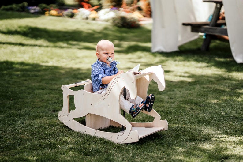 May include: A light-colored wooden rocking horse with a child seated in it, positioned on a grassy lawn. The horse features a cutout design and a seat. The child is wearing a blue shirt and light-colored pants.