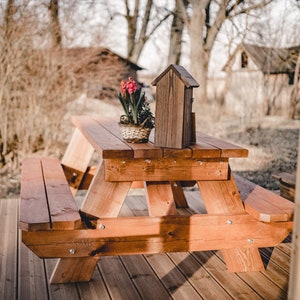 May include: A wooden picnic table with two benches, set on a wooden deck. A small wooden birdhouse and a potted plant with pink flowers are on the table.