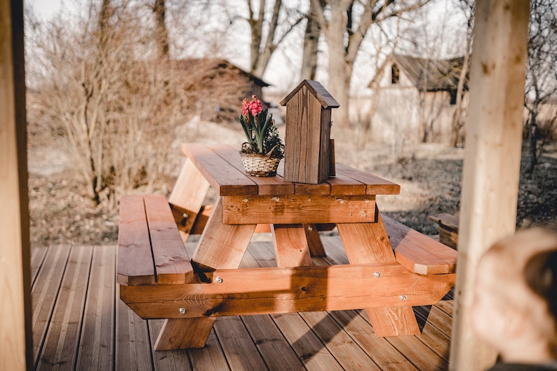 May include: A wooden picnic table with two benches, set on a wooden deck. A small wooden birdhouse and a potted plant with pink flowers are on the table.