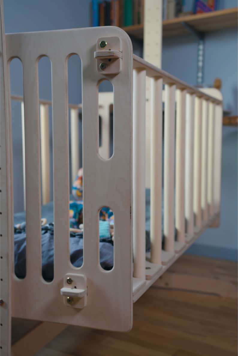 Puede incluir: Una cuna de madera clara con listones verticales y esquinas redondeadas. La cuna tiene un dise&ntilde;o sencillo y est&aacute; suspendida de una estructura de soporte. El interior contiene una s&aacute;bana azul y juguetes coloridos.