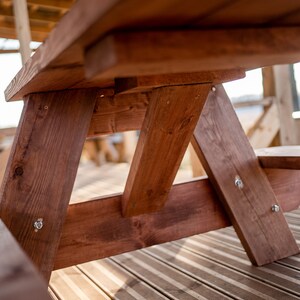 May include: Close-up of a wooden picnic table with a brown stain. The table is made of sturdy planks and has a simple, rustic design.