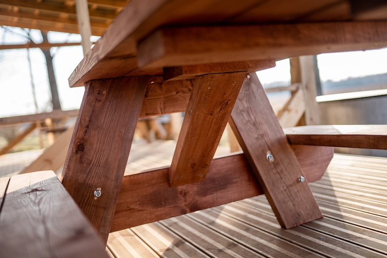 May include: Close-up of a wooden picnic table with a brown stain. The table is made of sturdy planks and has a simple, rustic design.