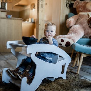 May include: A young child sits in a white wooden high chair with a black bowl on the tray. The child wears a blue striped shirt and jeans. A large teddy bear sits on a blue chair in the background.
