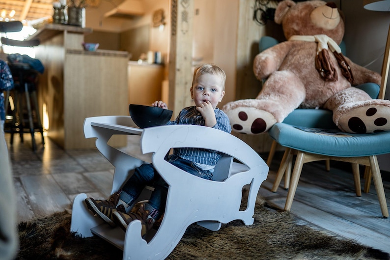 May include: A young child sits in a white wooden high chair with a black bowl on the tray. The child wears a blue striped shirt and jeans. A large teddy bear sits on a blue chair in the background.