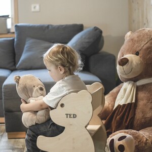 May include: A wooden bear-shaped chair with the name "TED" printed on it. A child is sitting in the chair, holding a stuffed bear. A large brown teddy bear is in the background. The chair is made of light-colored wood.