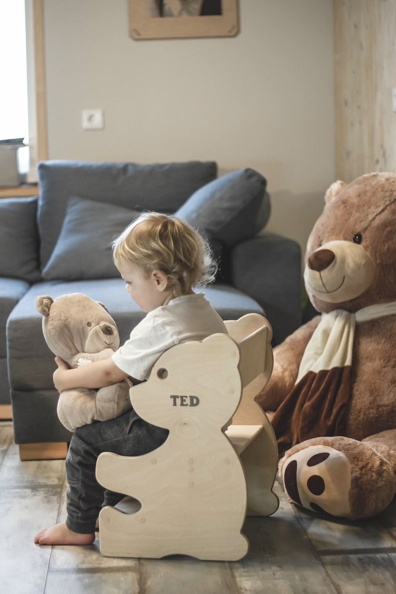 May include: A wooden bear-shaped chair with the name "TED" printed on it. A child is sitting in the chair, holding a stuffed bear. A large brown teddy bear is in the background. The chair is made of light-colored wood.