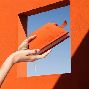 Puede incluir: Un cuaderno naranja con el texto "candie al aire" en la portada está siendo sostenido por una mano a través de una ventana en una pared naranja.