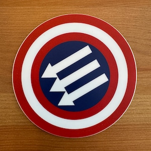 May include: A round sticker with a red, white, and blue design. The center of the sticker features a blue circle with three white arrows pointing upwards. The arrows are arranged in a triangular pattern.