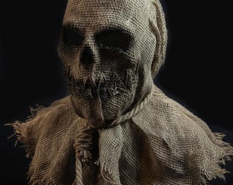 May include: A full-head mask crafted from coarse, tan-colored burlap fabric. The mask features deep, dark, hollow eye sockets and a stitched, jagged mouth opening. A thick, knotted rope is tied around the neck, with frayed edges at the base.