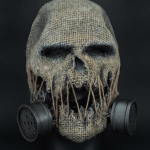 May include: A full-head mask crafted from coarse, tan-coloured burlap fabric, featuring deep eye sockets and a jagged, open mouth stitched with vertical twine. Two matte black, cylindrical gas mask filters are attached to the sides of the jaw.