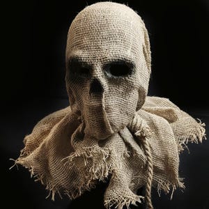 May include: A tan burlap mask featuring a skull-like structure with deep, dark eye sockets and a triangular nose opening. The mask includes a wide, frayed collar section and a thick, knotted rope cord tied around the neck area, creating a textured look.