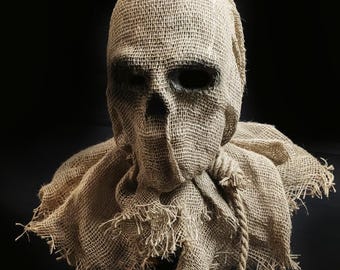 May include: A tan burlap mask featuring a skull-like structure with deep, dark eye sockets and a triangular nose opening. The mask includes a wide, frayed collar section and a thick, knotted rope cord tied around the neck area, creating a textured look.