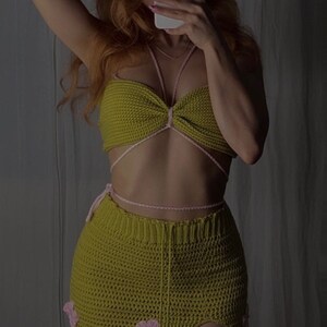 Puede incluir: Un conjunto de top y falda de crochet verde con detalles de flores rosas. El top tiene un estilo halter con un nudo en la parte delantera. La falda es una minifalda de cintura alta con una abertura lateral.