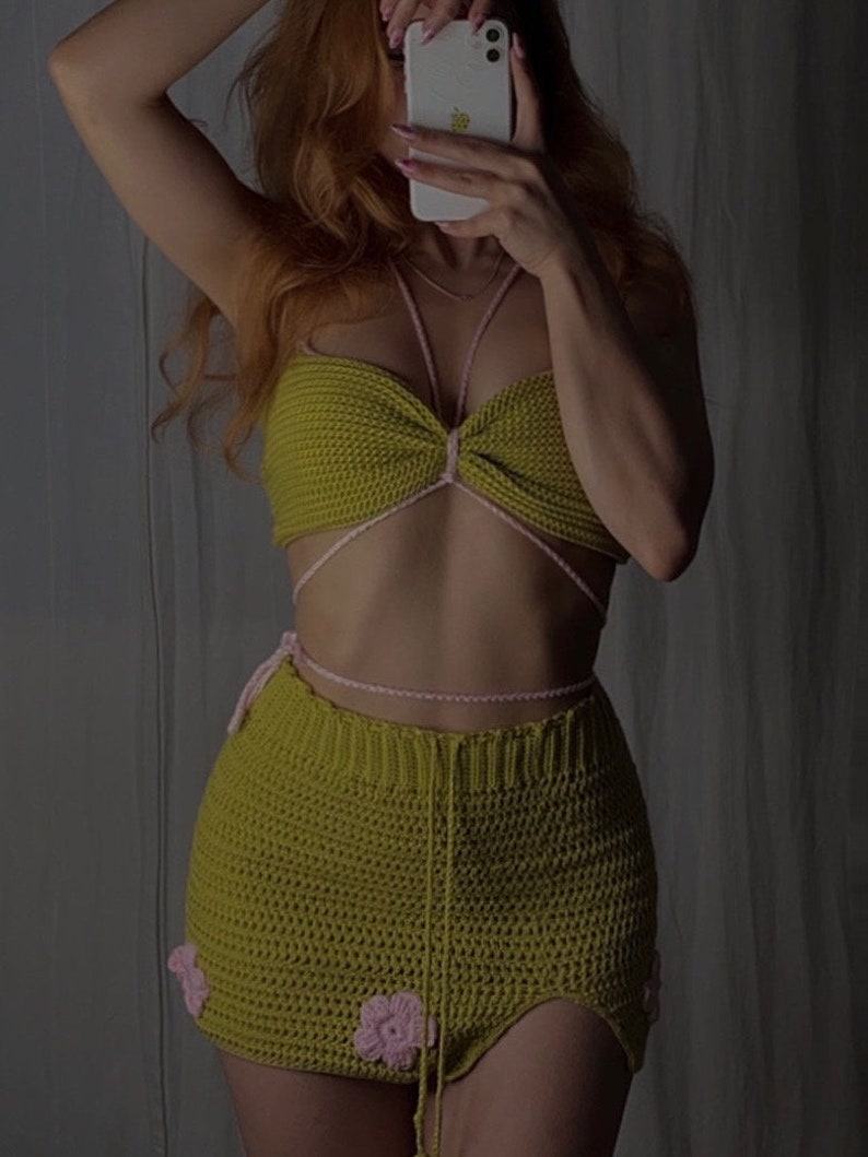 Puede incluir: Un conjunto de top y falda de crochet verde con detalles de flores rosas. El top tiene un estilo halter con un nudo en la parte delantera. La falda es una minifalda de cintura alta con una abertura lateral.