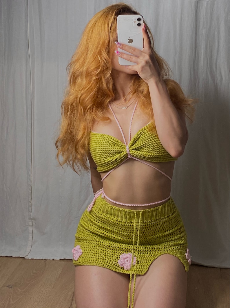 Puede incluir: Un conjunto de top y falda de crochet verde con detalles rosas. El top es de estilo halter con un nudo en la parte delantera. La falda es una minifalda con una abertura lateral y flores rosas como adorno.