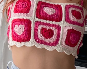 May include: A pink and white crocheted crop top with a heart pattern. The top has a scalloped hem and thin straps.