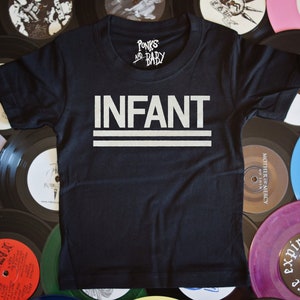 May include: Black t-shirt with white text that reads "Punks and Baby" and "INFANT" with two white lines underneath the word "INFANT". The shirt is laying on top of a stack of vinyl records.