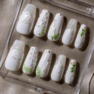 May include: White press-on nails with intricate designs. Some nails feature lace-like patterns, tiny silver beads, and delicate green leaf accents. The nails are arranged in a clear plastic case.