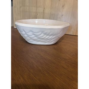 May include: A white ceramic bowl with a textured, wave-like pattern on the exterior. The bowl has a wide, oval shape and sits on a wooden surface. The interior of the bowl is smooth and white.