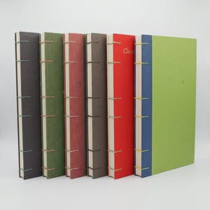 May include: A set of six hardcover books standing upright in a row. The covers are black, olive green, maroon, dark brown, red, and a combination of blue and lime green. Each book features exposed coptic stitching on the spine. The red book has the word Classic printed in gold on the spine.