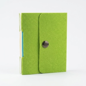 May include: A square lime green felt notebook with a rounded flap closure secured by a single dark metal snap button. The side of the notebook reveals thin, multicolored paper pages stacked vertically in shades of pink, yellow, and blue.