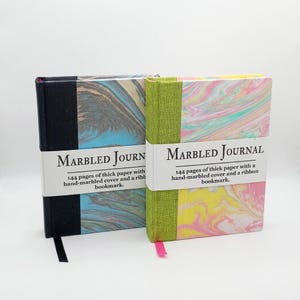 May include: Two journals with marbled covers. One journal has a black spine and the other has a green spine. Each journal has a white band with the words "MARBLED JOURNAL" and the description "144 pages of thick paper with a hand-marbled cover and a ribbon bookmark."