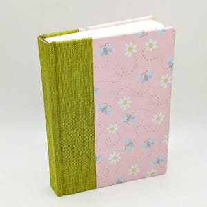 Könnte beinhalten: Ein grünes und rosa gebundenes Tagebuch mit einem Bienen- und Gänseblümchenmuster. Das Tagebuch hat einen grünen Leinenrücken.