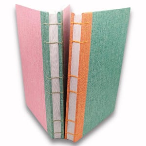 May include: Two handmade journals with colourful covers. One journal has a pink cover, the other has a green cover. The spines are hand-stitched with thread. The second journal has an orange spine. The pages are white.