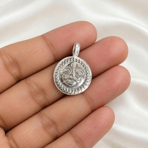 May include: A circular silver-toned metal pendant featuring a raised relief of a sun face with a mustache and a small vertical mark on the forehead. The outer edge of the pendant has a textured, rope-like border and a small loop at the top.