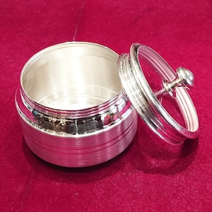 May include: A silver container with a lid. The container has a smooth, shiny finish and a slightly rounded shape. The lid has a small knob on top.