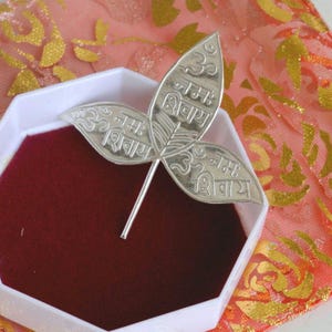 May include: A silver-toned metal pin shaped like a three-leaf clover or bilva leaf. Each of the three leaves is engraved with the Sanskrit text Om Namah Shivaya. The pin features a thin, straight metal stem extending from the base of the leaves.