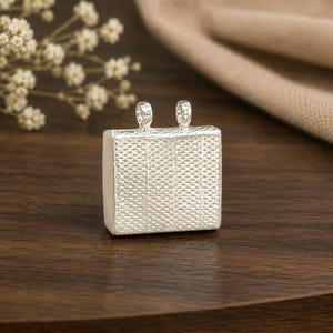 May include: A square silver pendant featuring a textured cross-hatch pattern on its front surface. The pendant has two small, vertical silver loops attached to the top edge, designed for threading a chain or cord through for jewelry making.