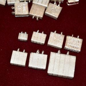 May include: A collection of silver-toned metal charms with intricate designs. Some charms have a textured surface, while others feature engraved patterns. The charms are arranged on a red surface.