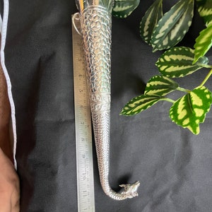 May include: A silver-coloured, ornate, horn-shaped object with a dragon's head at the curved end. The object has a textured surface and is approximately 30 cm long. A ruler is placed next to the object for scale.