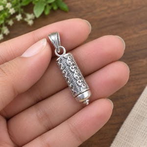 May include: A cylindrical silver pendant featuring an intricate embossed scrollwork pattern and a braided texture detail. The pendant includes a tapered bail at the top for attachment and a rounded, smooth cap at the bottom end of the metal cylinder.