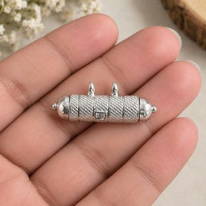 May include: A cylindrical silver pendant featuring a textured cross-hatched pattern on the main body, rounded end caps, and two small vertical loops on top. The metal surface is stamped with the hallmark 925, indicating sterling silver composition.